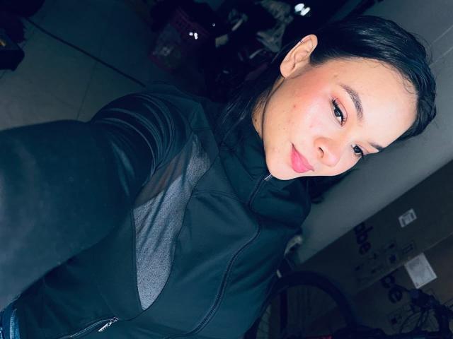 Fotografia de Yurimy, com pele clara e cabelo preto, vestindo uma jaqueta preta, tirando uma selfie. O volume dos genitais de Yurimy é sutilmente sugerido pelo corte da jaqueta. Sorrindo, com iluminação forte..