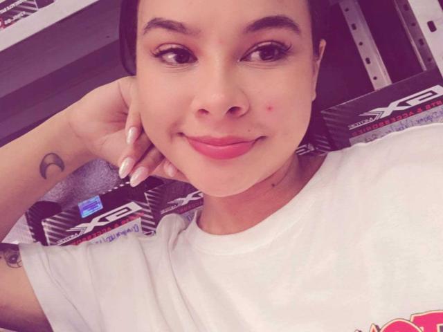 Close-up selfie of Yurimy, a young woman with light brown skin, dark hair, and pink lips, lying on a car seat with a black and white racing stripe pattern. She's smiling, hand behind head, showing a small crescent moon tattoo on her wrist. Her long nails are painted white. Fucking cute.