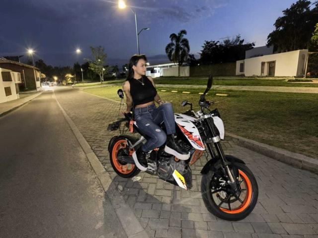 Yurimy, sentada en una motocicleta negra y blanca con llantas naranjas, vestida con una camiseta negra y jeans, al atardecer en un lugar iluminado. calle, farolas brillando en el fondo.