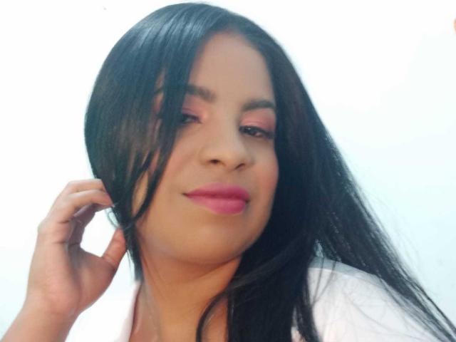 Close-up de Chloe Harrison, uma mulher com cabelos longos e pretos, sombra rosa e batom rosa, sorrindo levemente. A mão direita tocando o cabelo dela. Fundo branco. Incrivelmente atraente..