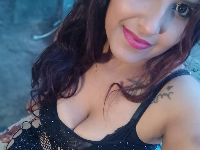 Close-up de DannaRodriguez69, uma mulher com cabelos longos e escuros, batom rosa e blusa de renda preta que revela bastante o colo. Tatuagem em seu ombro direito, iluminação com tons de azul, tensão sexual..