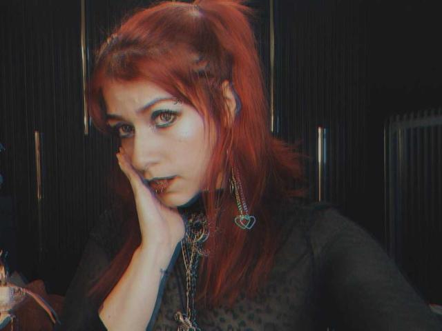 Fotografie von LolyYunno mit roten Haaren, viel Make-up und dunklem Lippenstift, die ihre Hand auf ihr Gesicht legt. Sie trägt ein schwarzes, durchsichtiges Oberteil und herzförmige Ohrringe. Verdammt intensiver Blick.