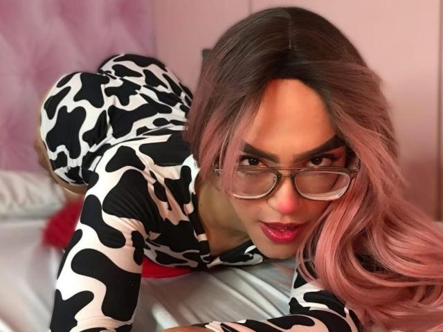 MelissaHotX69, com cabelo rosa comprido, vestido com estampa de vaca em preto e branco e óculos, inclina-se para frente em uma cama com um ar sedutor. expressão. Seus lábios vermelhos estão ligeiramente entreabertos. A tensão sexual é alta..