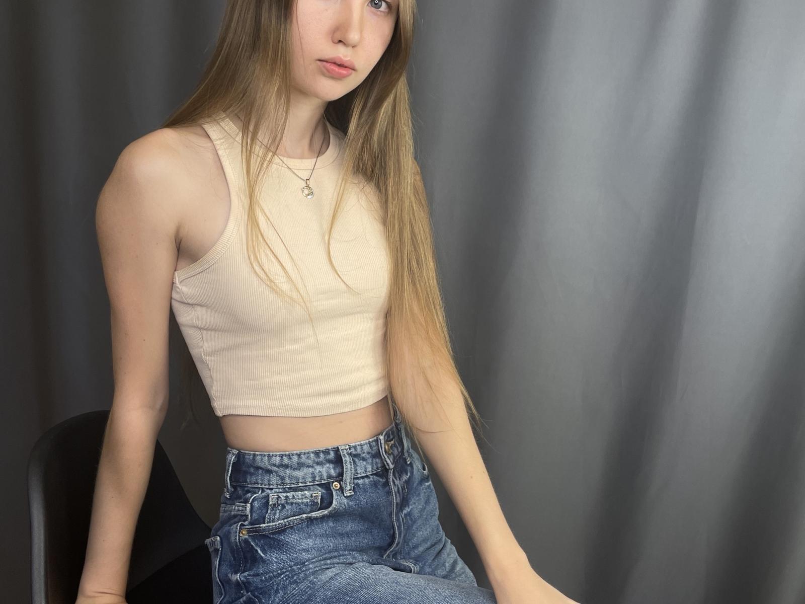 Jolene Ganther, com cabelos castanhos longos, vestindo uma regata branca justa com textura em tiras e calças jeans azuis de cintura alta, sentada contra uma cortina cinza escura.
tensão sexual, insinuações de ereção visíveis através do tecido..