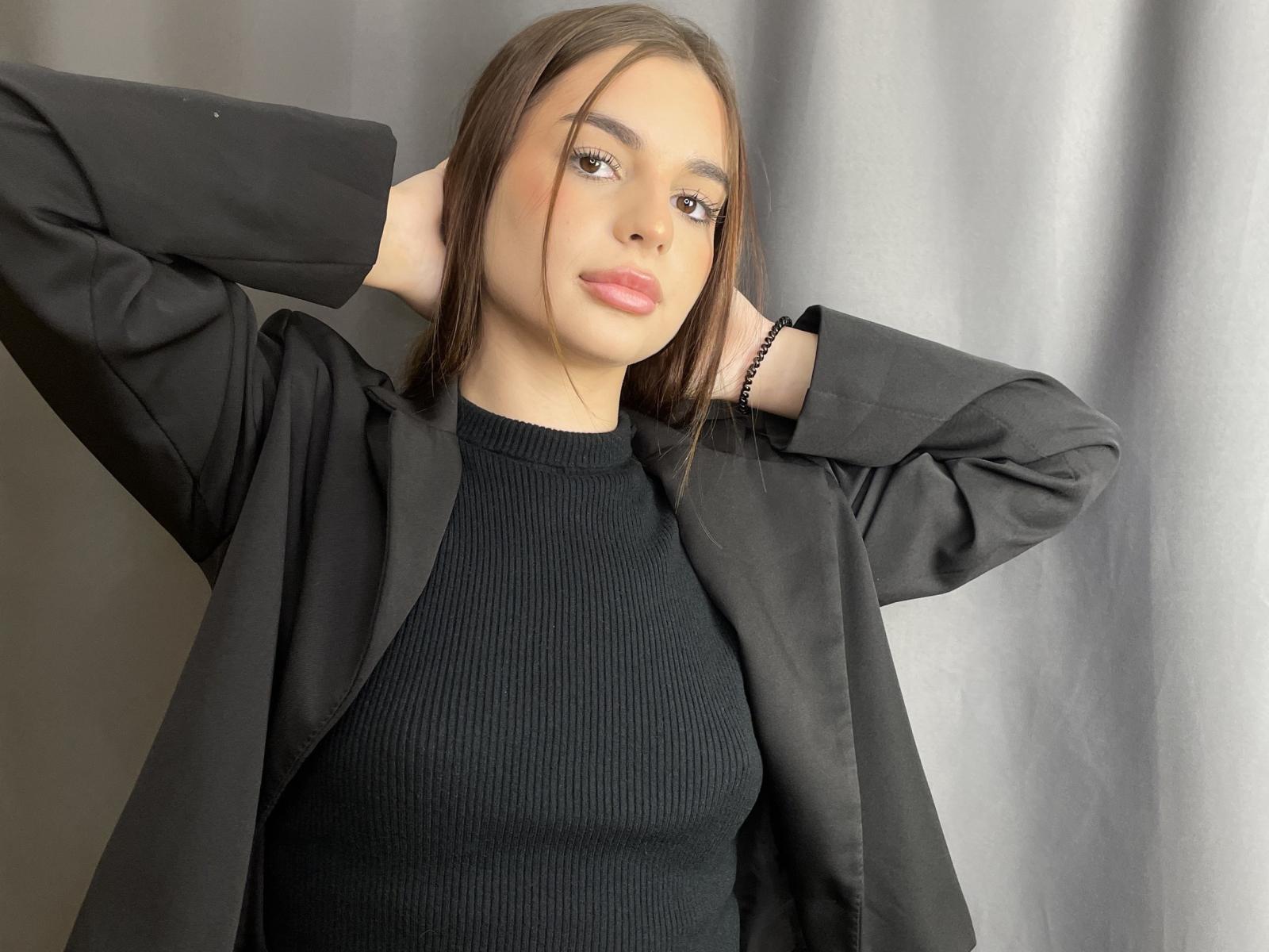 Roselee Vandiest, mit hellbrauner Haut und glattem, dunkelbraunem Haar, trägt einen schwarzen, gerippten Pullover und eine offene schwarze jacke, Arme hinter dem Kopf erhoben, neutraler Gesichtsausdruck, weiches, natürliches Licht..