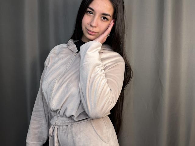 Sharmaine Tonnessen se tient devant un rideau gris, vêtue d'une chemise blanche à manches longues et à boutons, avec une ceinture. Elle touche son visage avec sa main droite, en souriant légèrement. Ses longs cheveux noirs tombent en cascade.
.