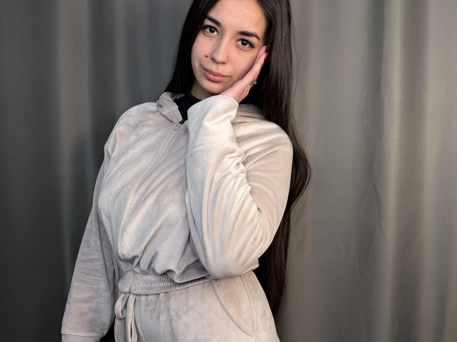 Sharmaine Tonnessen staat voor een grijze gordijn, gekleed in een witte, lang袖 shirt met knopen en een riem. Ze raakt haar gezicht aan met haar rechterhand en glimlacht zachtjes. Haar lange, zwarte haar valt in golven..