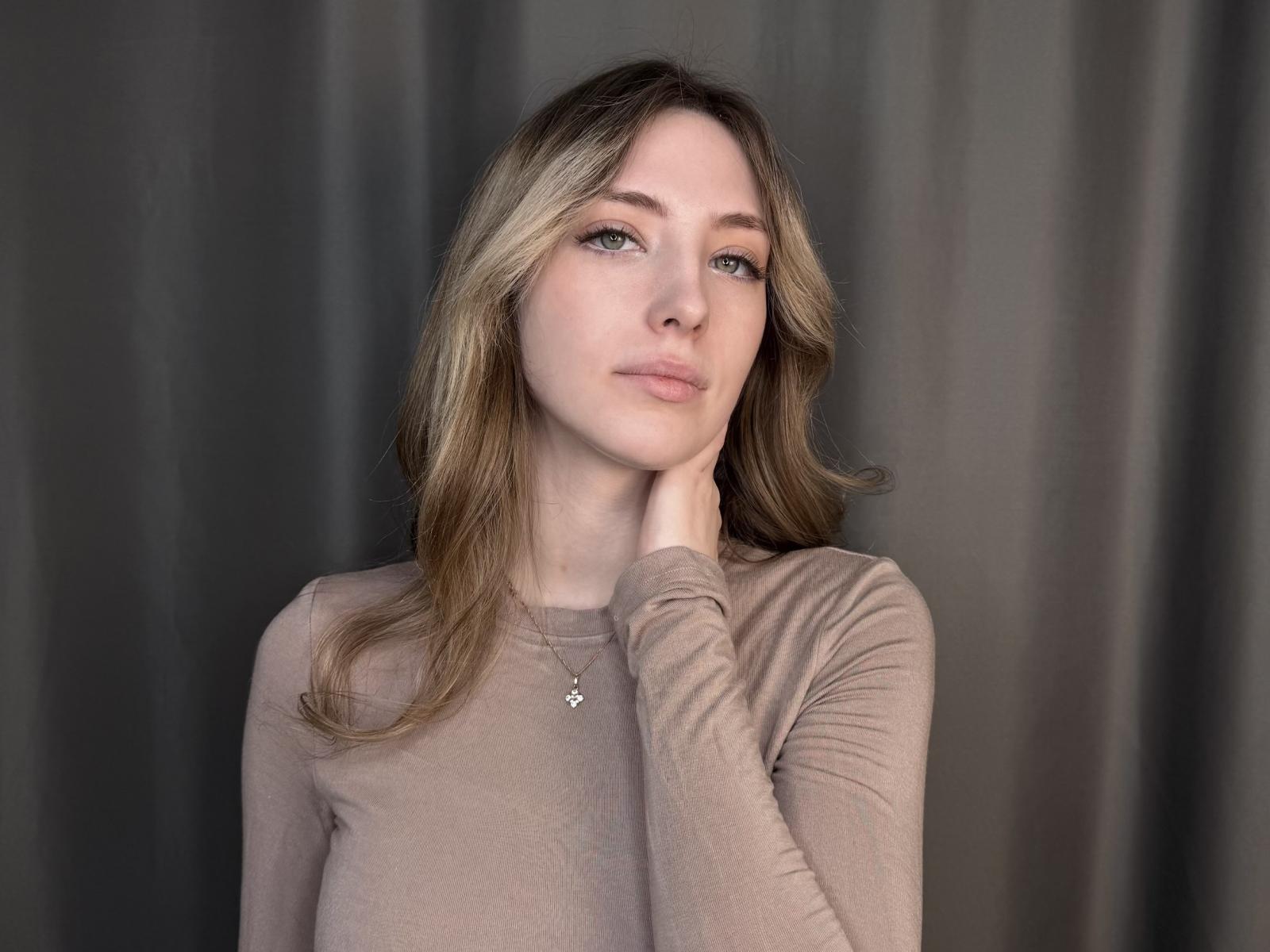 Photographie de JoseNoack, avec des cheveux bruns clairs, des yeux bleus et une expression neutre. Elle porte un haut marron transparent à manches longues, une main posée sur son cou, avec une légère tension sexuelle. Fond noir.