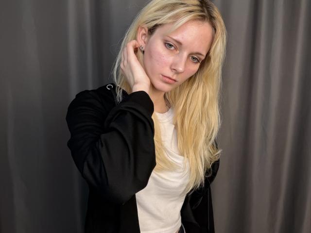 Photograph of DierdreLorenzano, a blonde woman with fair skin, wearing a white shirt and black jacket, touching her hair with a seductive, slightly distant gaze, gray curtain background.