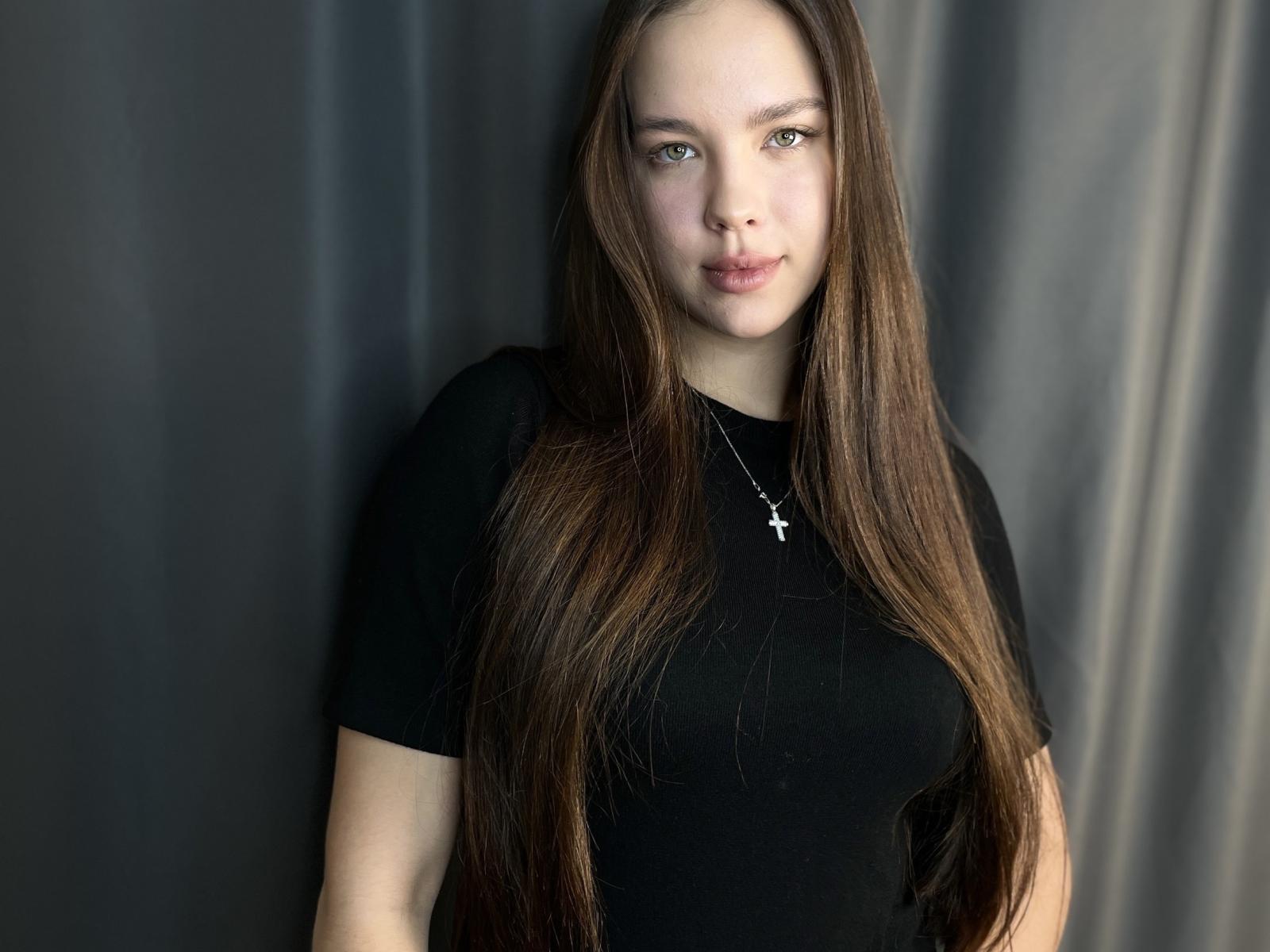 ShilaBrunetti staat voor een donkere, bedekte achtergrond, gekleed in een zwart T-shirt, met lang, bruin haar dat over haar valt. schouders, lichte make-up, serieuze uitdrukking, subtiele schaduwen, lichte blush, zachte belichting.