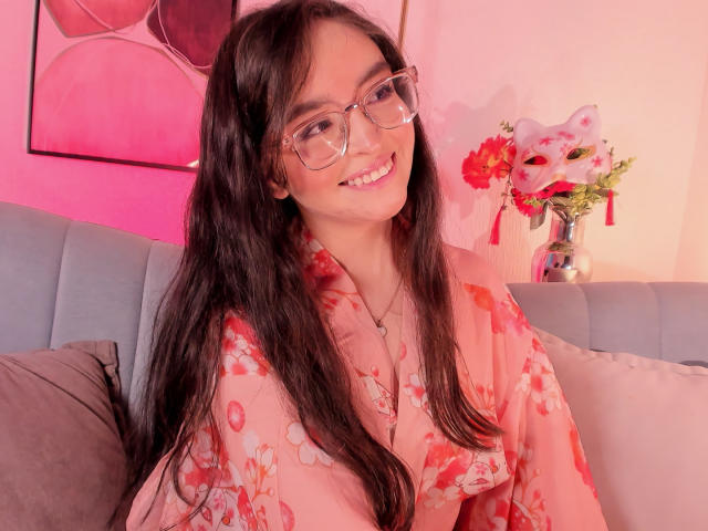 EvaMcAllister, een schattige Aziatische vrouw met lang zwart haar en een bril, glimlacht verleidelijk terwijl ze een roze badjas met bloemenprint draagt. Roze verlichting, grijze bank, rode bloemen op de achtergrond. Verdomd aantrekkelijk..