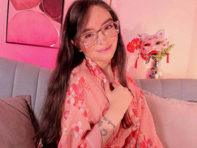 Eva McAllister mit langem, schwarzem Haar, die eine Brille trägt und einen pinken Kimono mit Blumenmuster anhat, sitzt auf einem grauen Sofa, lächelt und hat eine Hand erhoben. in der Nähe des Kinns, roter und pinker Hintergrund, Blumen im Hintergrund.