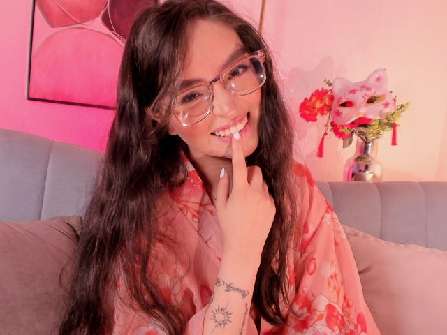 EvaMcAllister, una atractiva mujer asiática con cabello negro largo y gafas, sonríe de manera seductora, mordiéndose el dedo. Ella lleva un kimono rojo con estampado floral, sentada en un sofá rosa. Flores vibrantes en el fondo..