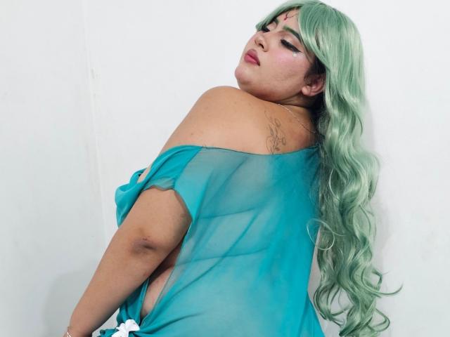 Fotografie von GraceYuly, mit dem Rücken zur Kamera, die über die Schulter schaut. Sie hat langes, welliges, grünes Haar und trägt ein durchsichtiges, blaues Oberteil, das ihren Rücken und ihre Schultern freilegt. Sexuelle Spannung, verführerischer Blick.