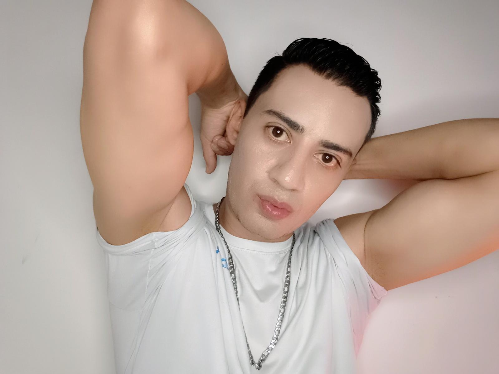 Photograph of LucianoZ, lying on a white surface, arms raised behind head, brown eyes, short black hair, white sleeveless shirt, chain necklace, slight smile, soft light, relaxed pose, sexy vibe.