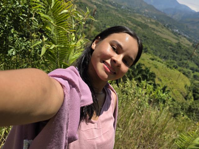 Yurimy se toma un selfie al aire libre, inclinándose ligeramente hacia adelante y con una pequeña sonrisa. Ella tiene el pelo largo y negro, piel morena, y lleva una camisa rosa. La luz del sol ilumina su rostro y el fondo verde del bosque..