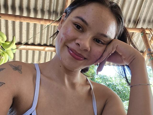 Close-up of Yurimy with a sexy smile, dark hair, and small tattoos on her shoulder. She's wearing a white strap top, hand on cheek, under a corrugated metal roof. Bright light.