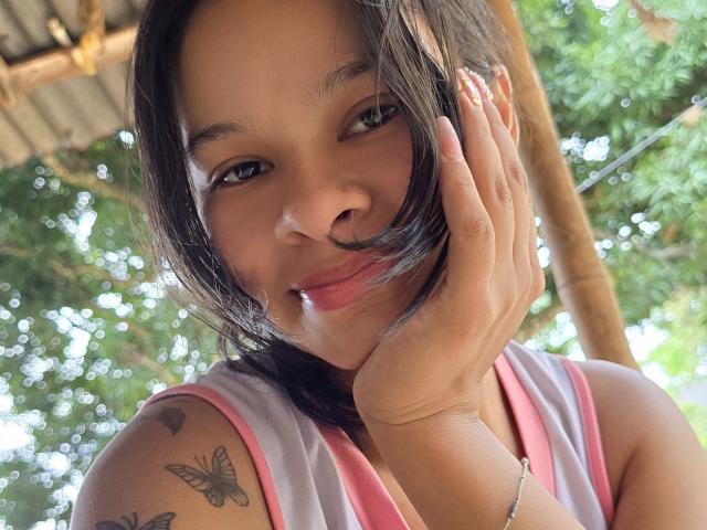 Close-up de Yurimy, uma mulher de pele morena e cabelo preto, sorrindo de forma sedutora, com a mão tocando sua bochecha, e uma borboleta preta. Tatuagens nos ombros, vestindo uma blusa rosa sem mangas, fundo de uma floresta verde..