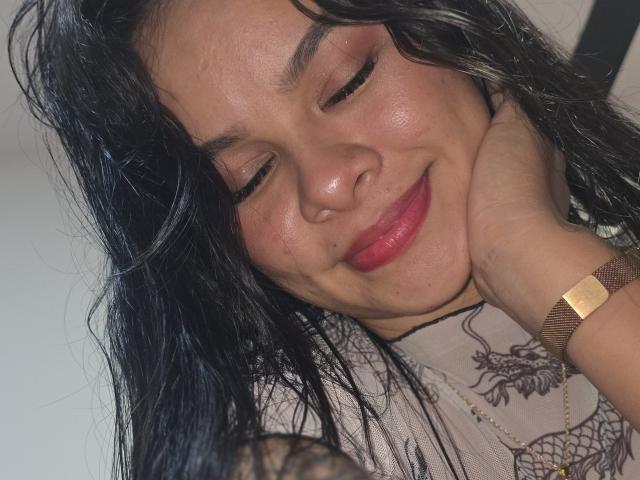 Nahaufnahme von Yurimy, einer Frau mit dunklen, welligen Haaren, rotem Lippenstift und einer goldenen Uhr am Handgelenk, die mit ihren Augen lächelt. geschlossen. Ihre Hand ruht auf ihrer Wange, was eine entspannte und verführerische Ausstrahlung vermittelt..