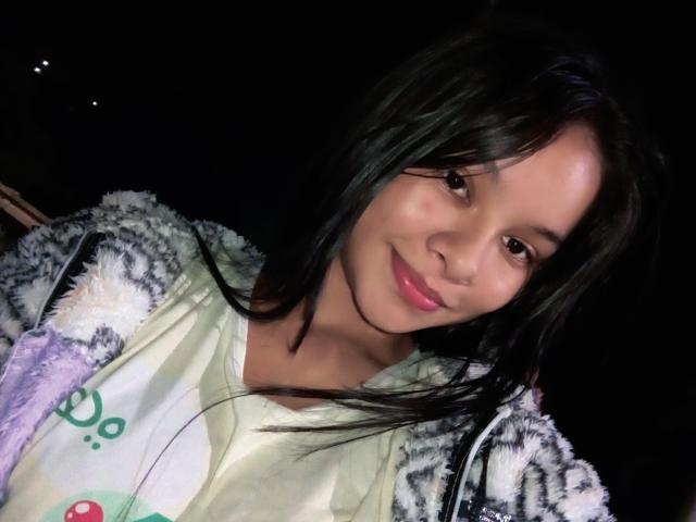 Photograph of Yurimy, a young woman with long black hair, smiling slightly, wearing a white shirt and fuzzy black-and-white patterned jacket, illuminated by a bright light source. Fucking sexy.