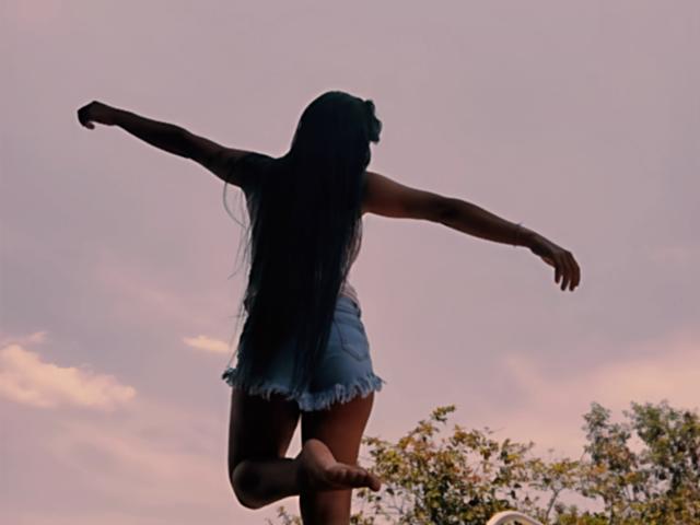 Fotografía en silueta de Yurimy, de espaldas a la cámara, con los brazos extendidos, vistiendo unos pantalones cortos vaqueros desgastados, y con el pelo largo y oscuro. De pie sobre una pierna, contra un cielo de atardecer rosa y morado con las copas de los árboles. Postura increíblemente atractiva..