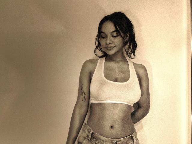 Sepia-toned photo of Yurimy with wet hair, wearing a white sports bra and jeans, standing with hands behind her back, slight smile, visible tattoo on right arm, casual pose.