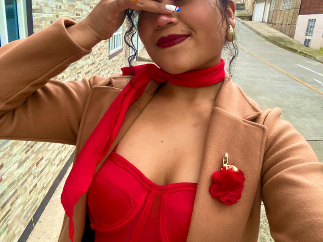 Fotografía de SophieSonyas con piel de color marrón medio, cabello rizado oscuro, lápiz labial rojo y una bufanda roja, vistiendo un blazer color beige. sobre una blusa roja con escote, con la mano cubriendo sus ojos. Fondo de calle.