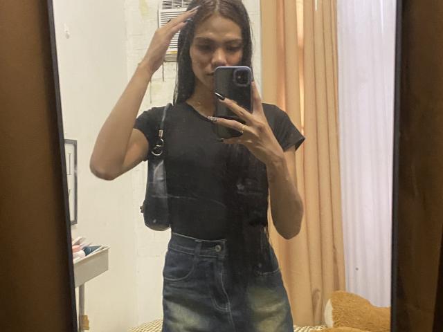 CassandraWise macht ein Selfie vor einem Spiegel, trägt ein enges schwarzes T-Shirt und eine Jeans mit hoher Taille, hat eine Hand im Haar und hält ein telefon, sinnliche Anspannung, gebräunte Haut, glattes schwarzes Haar, Zimmer mit beige farbenen Vorhängen.