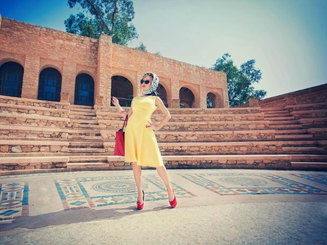 Shezel, vestida con un vestido amarillo brillante, tacones rojos y un bolso rojo, está de pie con confianza, de espaldas a la cámara. frente a antiguos escalones y arcos de ladrillo, bajo un cielo azul..