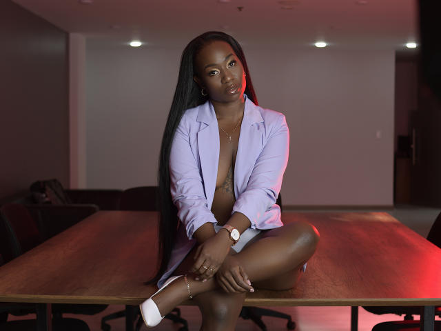 TamaraaJonss est assise sur une table en bois, vêtue d'une chemise blanche déboutonnée, laissant entrevoir sa poitrine, avec de longues tresses. montre-bracelet.
la lumière rouge projette une lueur séduisante. Elle regarde directement l'appareil photo..