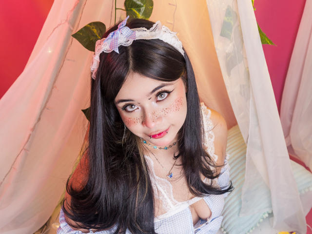 AmaraKazumi, with black hair and freckles, wears a white lace headband and dress with a low-cut neckline, showing cleavage. Soft light filters through white curtains, creating a sensual atmosphere.