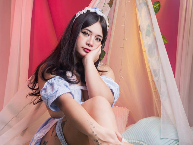 AmaraKazumi with long black hair, wearing a white lace headband and blue checkered off-shoulder top, resting her chin on her hand, sitting by a sunlit window with pink and white curtains. Fucking sexy.
