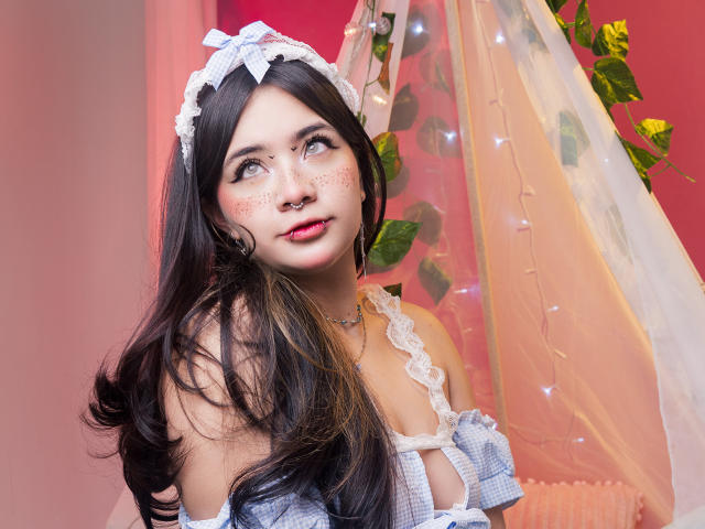 AmaraKazumi, con cabello negro largo y pecas, lleva un traje de sirvienta blanco de encaje y una diadema, mirando hacia arriba. El fondo presenta una pared rosa, cortinas translúcidas y flores amarillas. Qué lindo/a..