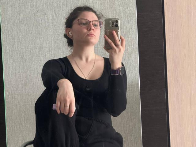 CariWannarka, con i capelli castano ricci, gli occhiali e una maglia nera a maniche lunghe, che tiene un telefono nella mano destra, è in posizione eretta. in primo piano, indossa una collana d'argento, guarda la fotocamera.