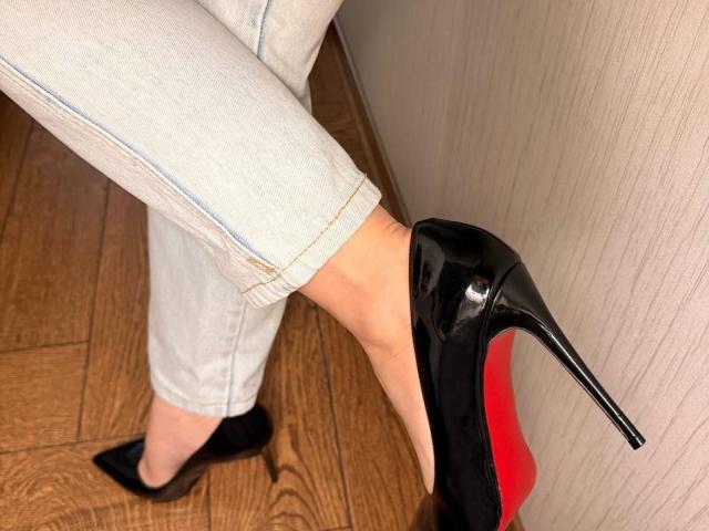 Close-up of KaiaLein's legs wearing white pants and black high heels with red soles, one foot slightly raised against a textured wall. Wooden floor, black slip-on shoe visible.