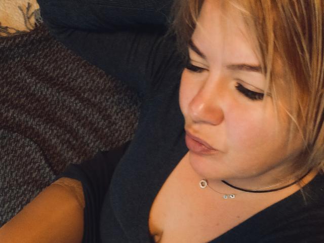 Primer plano de CurlyDreams, una mujer de piel clara con cabello rubio, labios carnosos y un collar negro. Ella lleva una blusa negra y mira hacia abajo con una expresión ligeramente seductora. Sus ojos están medio cerrados.
.