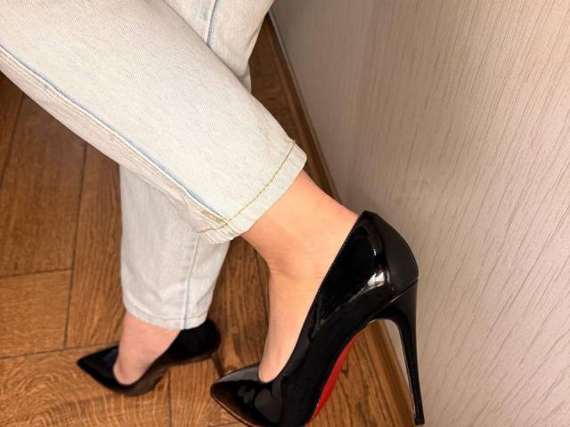 Close-up of KaiaLein's feet wearing black patent leather stiletto heels with red soles, standing on wooden flooring. KaiaLein's legs are in white pants, pressing against a textured wall. Fucking sexy.