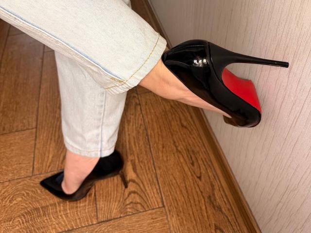 Close-up of KaiaLein's right leg in white pants, wearing black high heels with red soles, stepping on wooden floor. Shiny black shoes, wooden texture, strong sexual tension, provocative stance.