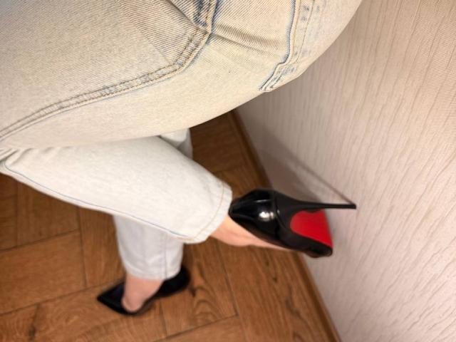 Close-up of KaiaLein's legs in white pants and black high heels with red soles, standing on wooden floor, leaning against white textured wall. Sexual tension implied by the angle.