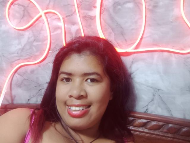 Close-up of MariahSquirting69 with medium brown skin, black hair, and red lipstick, smiling against a bright red neon sign on a marble wall. Visible shoulder strap, sexual tension, seductive gaze.