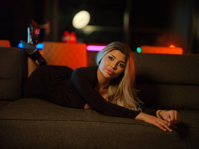 MoniqueCelestee ligt verleidelijk op een donkerbruine bank, gekleed in een strak zwart top, met haar lange blonde haar dat over haar schouders valt. haar schouder. Helder neonlicht in de achtergrond creëert een prikkelende sfeer..