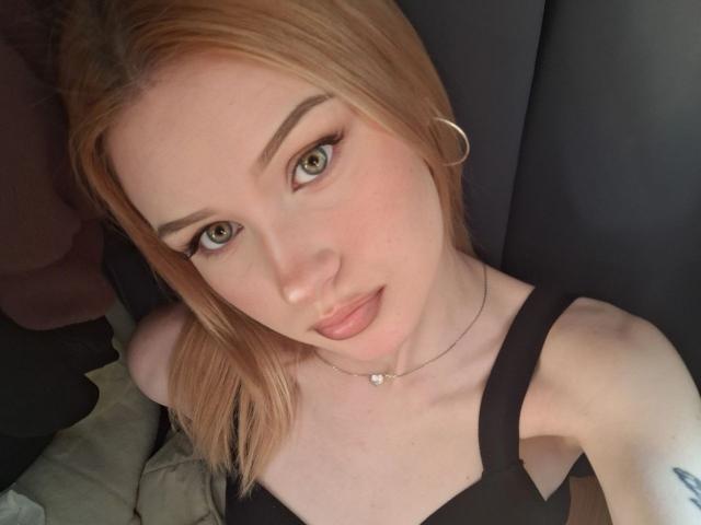 Close-up selfie of RoseUty with light brown hair, green eyes, and full lips. She's wearing a black tank top, hoop earrings, and a delicate necklace. Fucking hot, direct gaze.