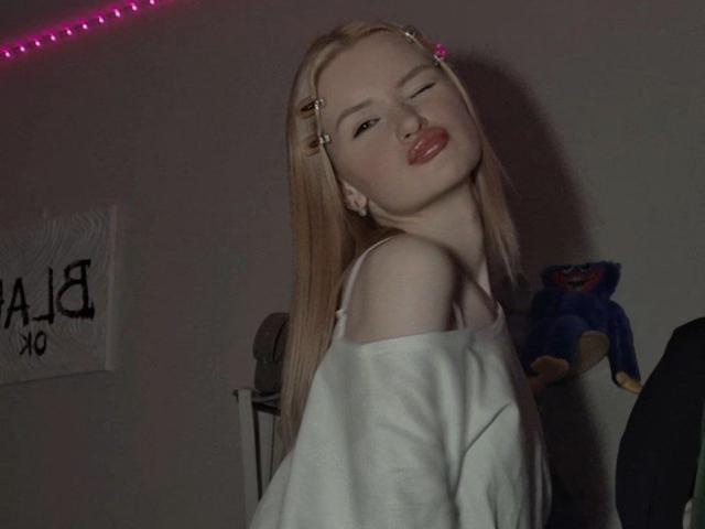 RoseUty, avec ses cheveux blonds et ses barrettes, porte un haut blanc à une épaule, et plisse ses lèvres dans une pose séduisante, avec des lumières LED roses. des lumières en arrière-plan, et une affiche partiellement visible. Extrêmement sexy..