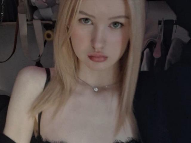Close-up van RoseUty met blond haar, blauwe ogen en roze lippen, die een zwarte top met bandjes draagt en een ketting met een klein hangertje. Ze kijkt recht in de camera met een licht opgewonden uitdrukking..
