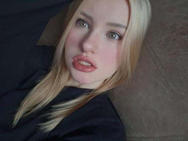 Close-up selfie of RoseUty with blonde hair, pale skin, and blue eyes. She has full, slightly parted pink lips. She's wearing a black top, leaning back on a dark surface. Fucking sexy.