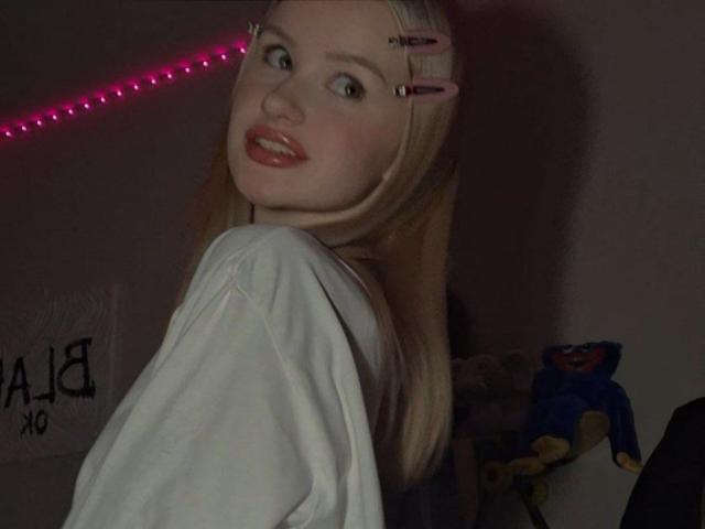 RoseUty, a blonde woman with pink hairpins, gazes back over her shoulder with a seductive smile. She wears a white shirt, illuminated by pink LED lights. Dark, moody background.