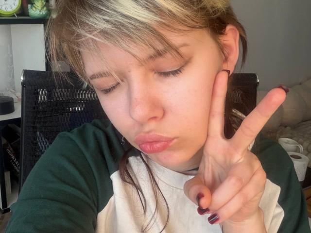 Close-up photo of LaurenceCloud with light brown hair, making a peace sign with their right hand, eyes closed, wearing a black and white raglan shirt, dark nail polish, soft natural light.