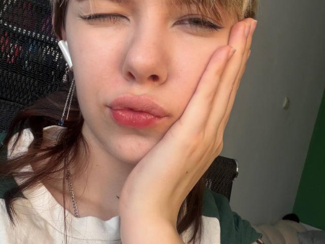Fotografía de primer plano de LaurenceCloud con piel clara, cabello castaño y labios rojos, con la mano apoyada en la mejilla, los ojos medio cerrados. tensión sexual sutil, luz natural, camisa blanca, pose informal.