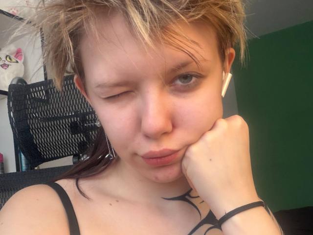 Gros plan de LaurenceCloud, avec des cheveux blonds en bataille, qui cligne de l'œil et pose sa joue sur sa main. LaurenceCloud a la peau claire, de petits tatouages et une bandoulière noire qui traverse sa poitrine. Tellement mignon..