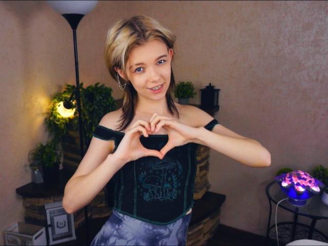 LaurenceCloud, a slim young woman with short blonde hair, makes a heart shape with her hands over a black off-shoulder top. She's smiling, standing indoors with dim lighting and plants.